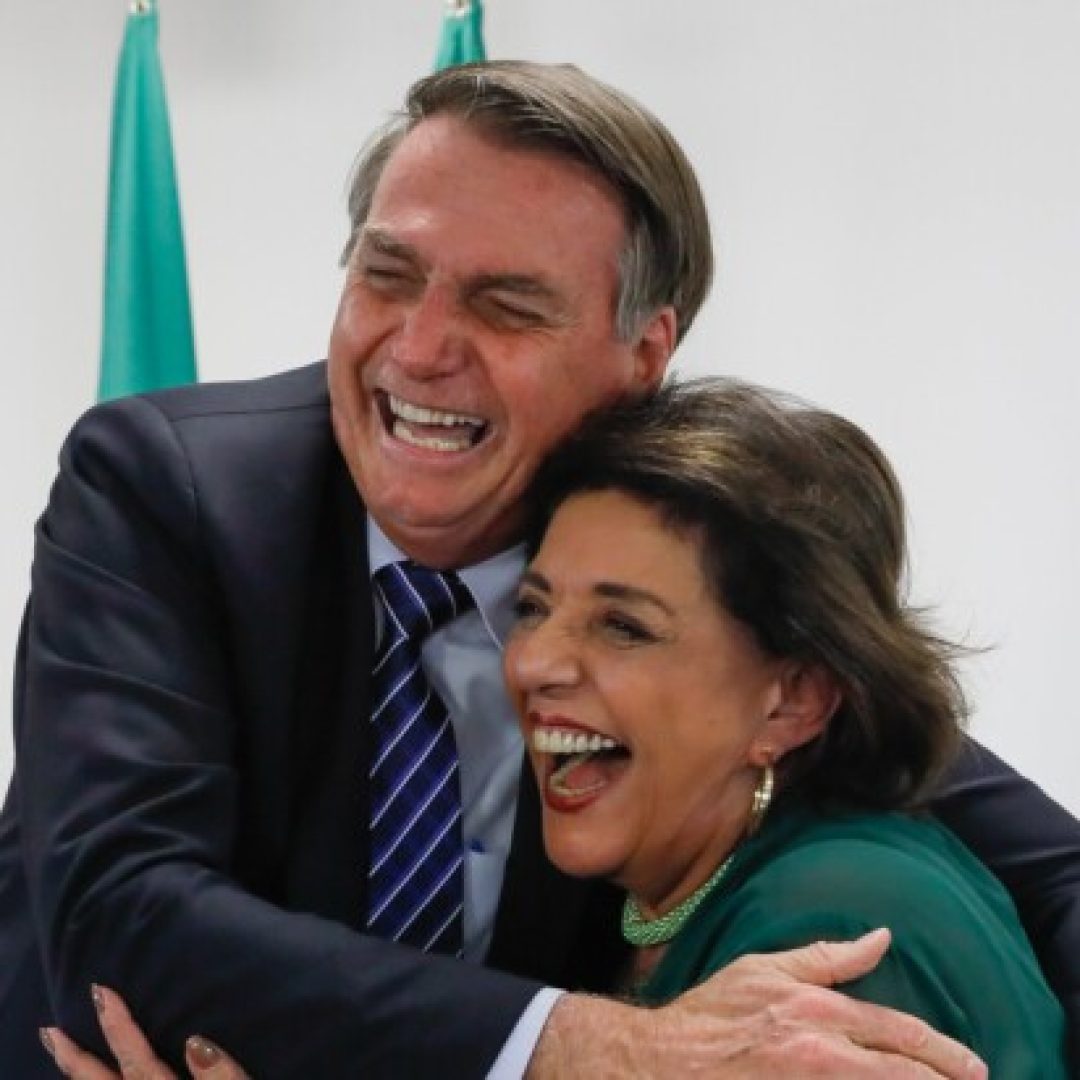 O presidente Jair Bolsonaro e a jornallista Leda Nagle se abraçando e rindo.